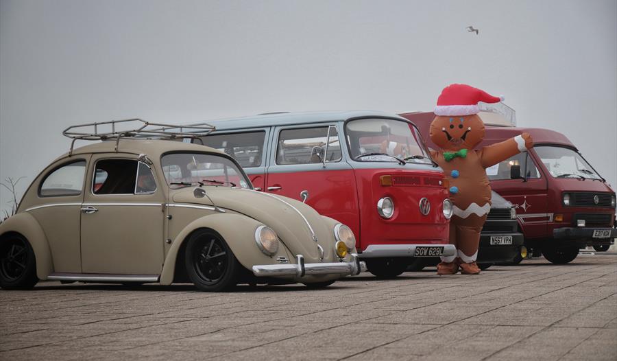four vintage volkswagens, with beetle in the foreground and three campervans behind. A giant gingerbread character is standing next to them.