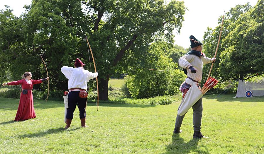 Two men and women extending bows and shooting archery targets on lawn. They are in medieval dress.