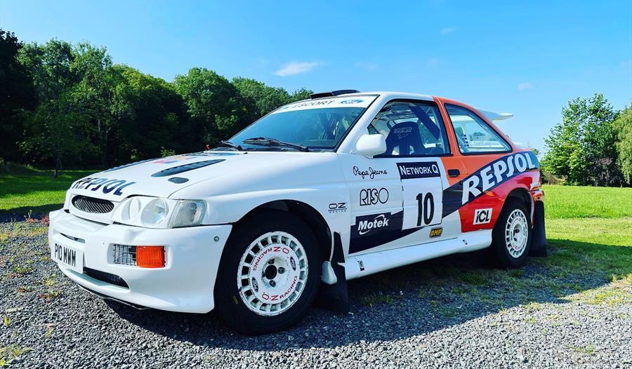 a white rally car with racing stickers over it, parked on gravel with green lawn and blue sky in background.