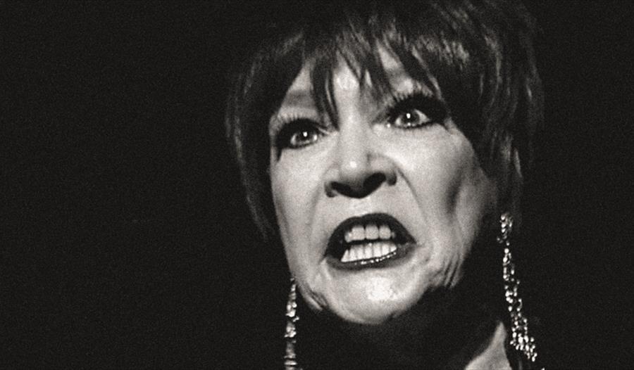 a black and white photograph, close up portrait of a woman's face with completely black background. Woman has dark lipstick and bears her teeth in an