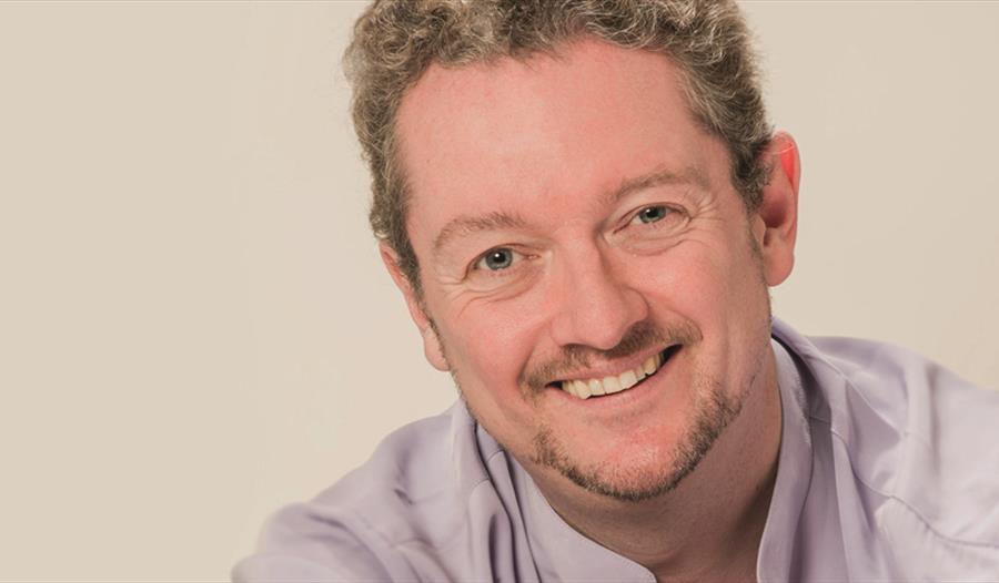 a portrait of a white man, smiling, just visible lilac shirt against a light plain background.