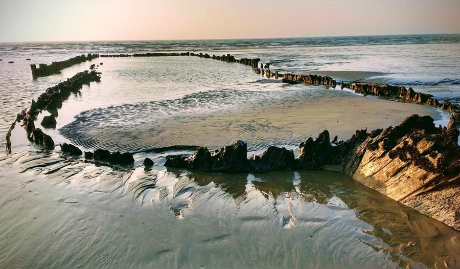 a photograph of the wreck the amsterdam. shows wet sand at very low tide with outline of boat emerging from it.