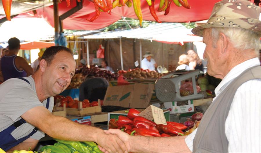 A market seller shaking hand across his veg stall with a man wearing a hat.