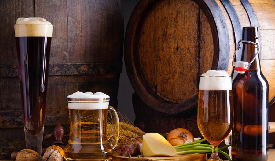 Photograph of three glasses of ales, with wooden barrel in background, surrounded by vegetable and cheese produce.