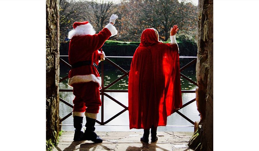 backs of father christmas and woman in red cloak waving out of castle archway.