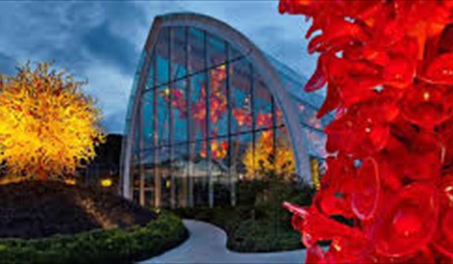 photographs shows cotemporary glass house at dusk with coloured glass sculptures in foreground, looking like trees.
