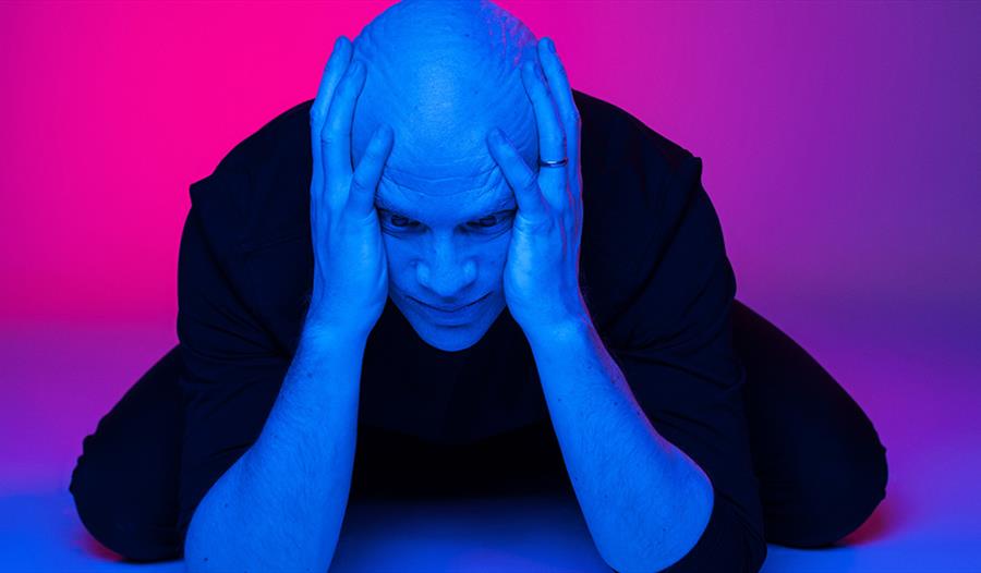 Photograph of a bald white man with his hands clasping the side of his head. the background and lighting is a hue of blue and pink.