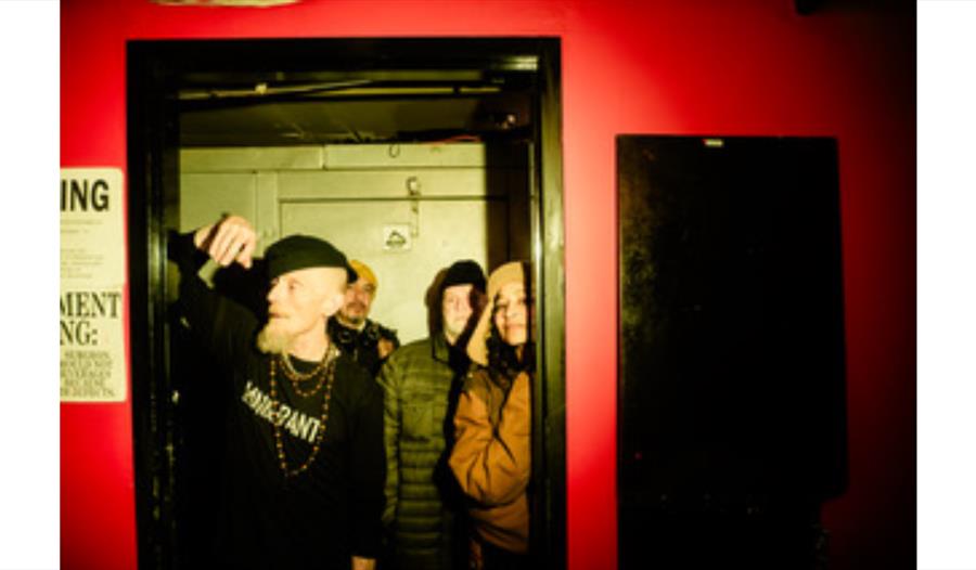 A dimly lit indoor scene with four people standing inside a doorway framed by a black doorframe. The surrounding wall is painted bright red, and there