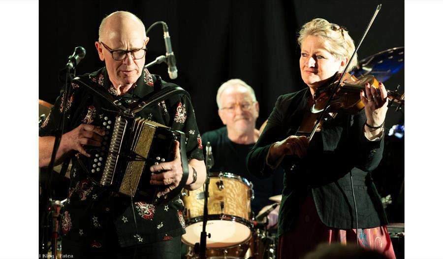 Three musicians perform on stage under warm lighting against a dark backdrop. On the left, an older man wearing glasses and a patterned black shirt pl