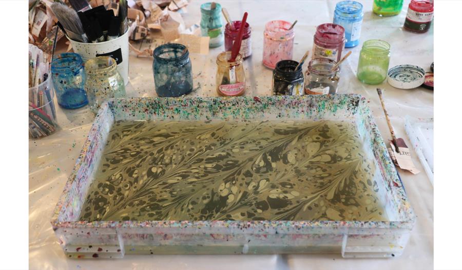 Tray of marbled water surrounded by jars of paint and tools in a paper marbling workshop.