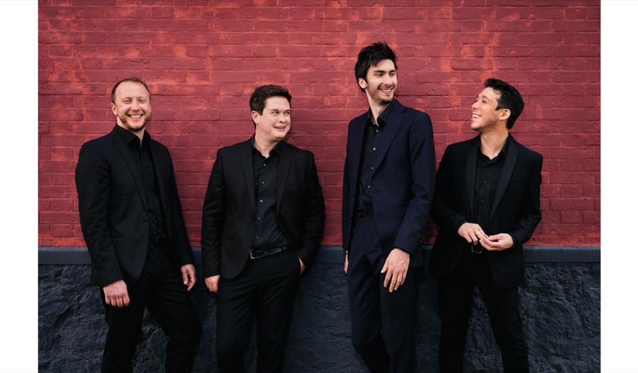 Van Kuijk String Quartet members in formal attire standing together against a red brick wall.