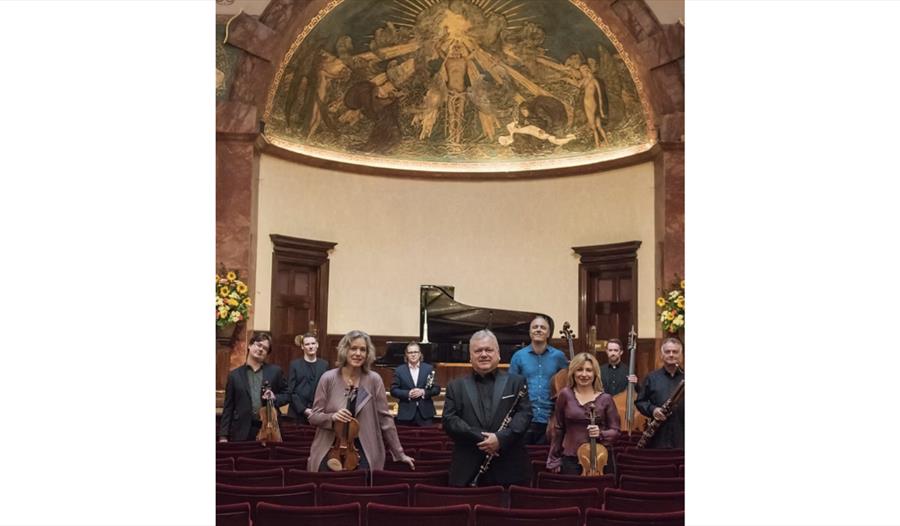 Wigmore Soloists ensemble posed with string, wind and piano instruments inside an ornate concert hall beneath a historic mural.