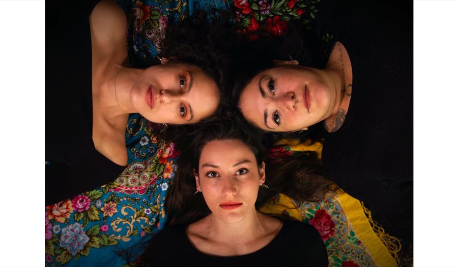 Promotional image of Alkanna Graeca, a Greek vocal trio, with three performers lying head to head on patterned fabrics.