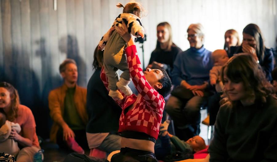 A performer in a red checkered top lifts a baby during "A Dance for Us" at Hastings Library, surrounded by families.