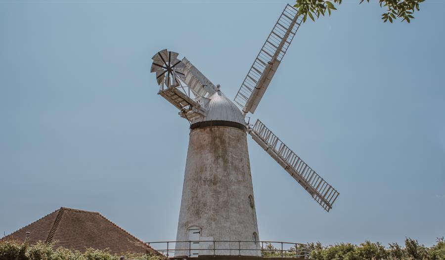 Stone Cross Windmill viewed from below, framed by leafy branches against a blue sky, with sails extended above a grassy area.