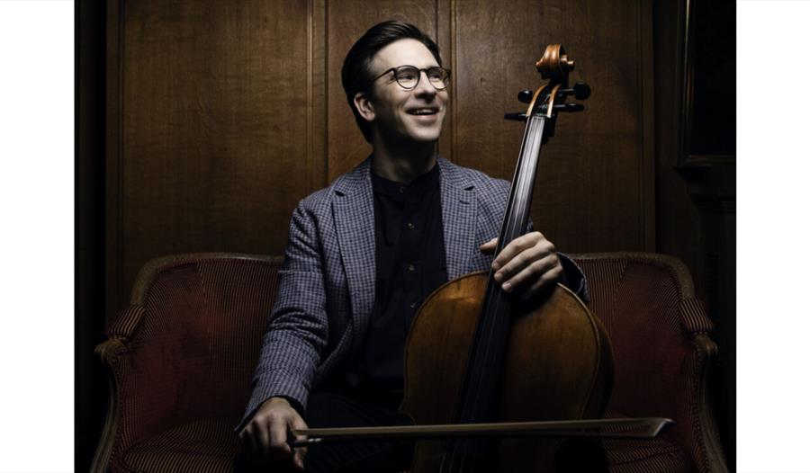 Promotional portrait for Guy Johnson and Friends featuring a cellist seated with cello in an elegant wood-panelled setting