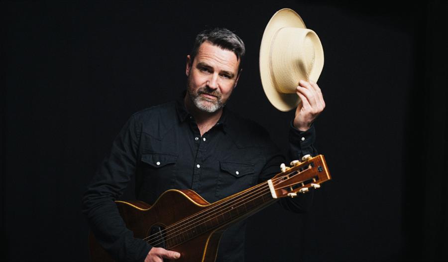 Promotional image for Martin Harley in The Shop – Late Show, featuring a musician with an acoustic guitar and hat.