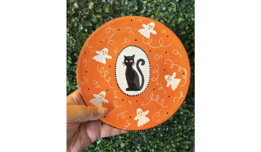 Hand-painted Halloween plate featuring a black cat, friendly ghosts and orange seasonal design at a pottery workshop.