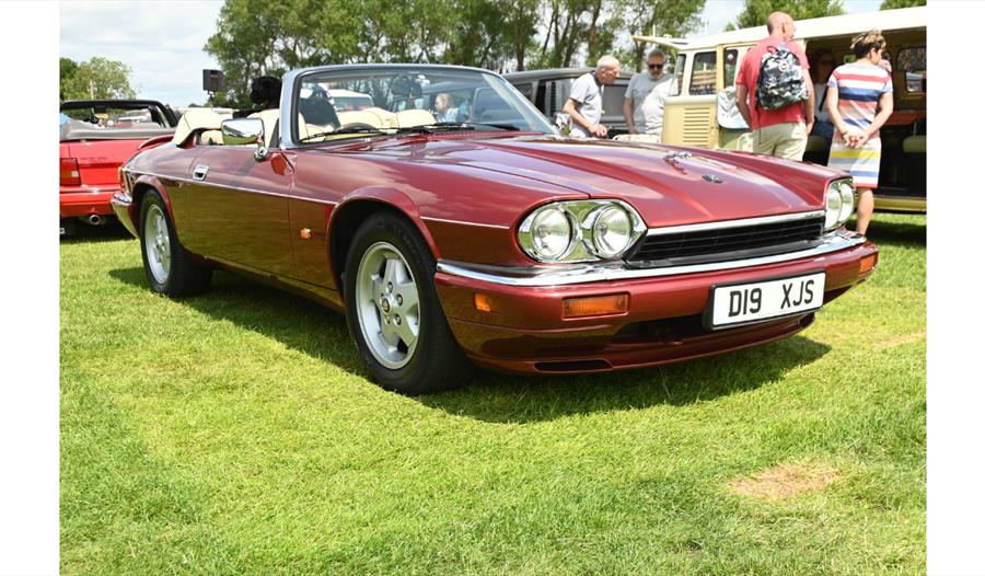 Red classic convertible sports car parked on grass at an outdoor car show, with people and other vintage vehicles in the background.