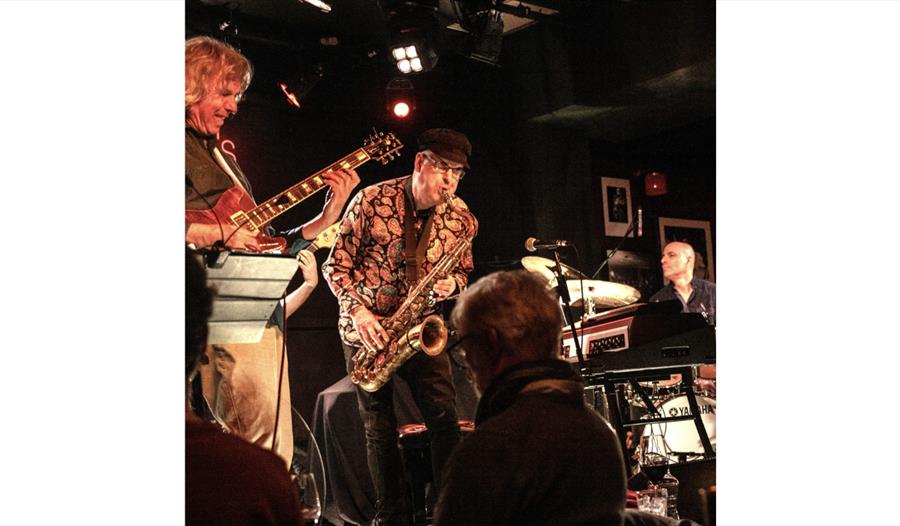 Soft Machine performing live at HJF ’26, with saxophone, guitar and keyboards on a warmly lit stage.