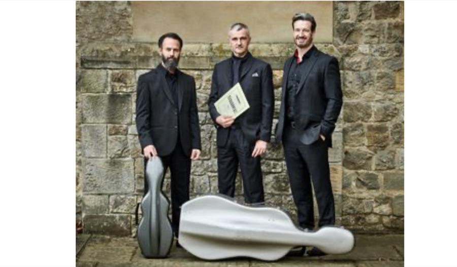 Tier3 and Friends performers in formal concert dress standing beside instrument cases against a stone wall backdrop