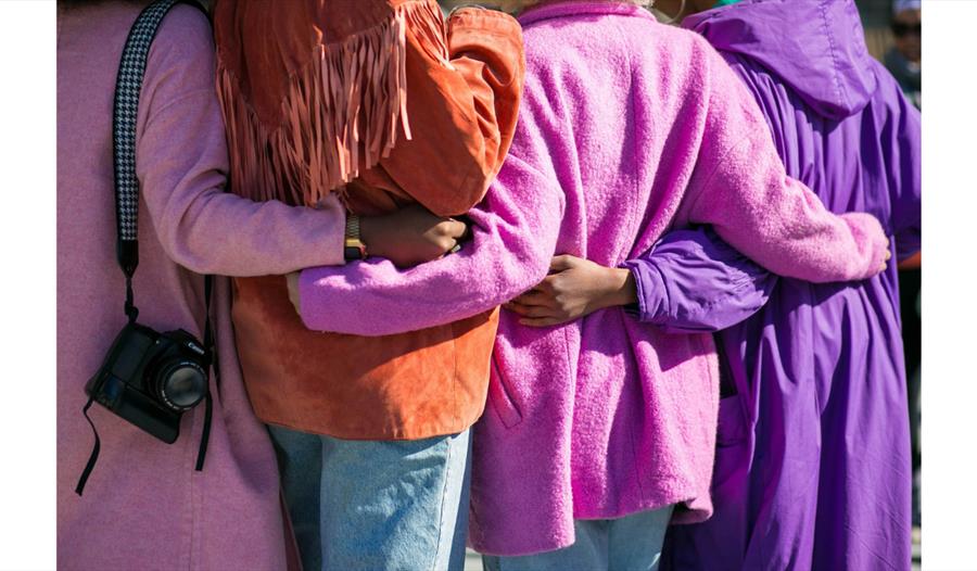 Group of women standing arm-in-arm in a supportive circle, representing connection, reflection and wellbeing.