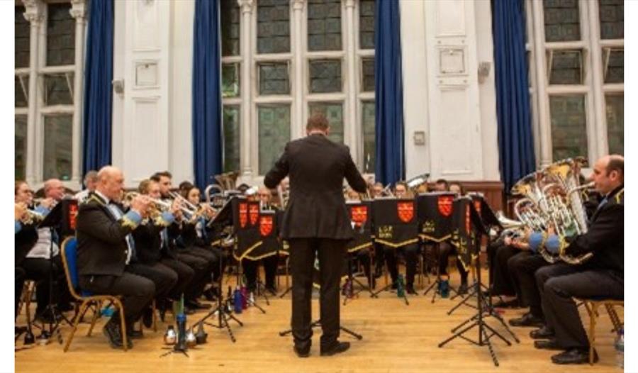 Lewes Brass band performs on stage with a conductor, arranged in rows inside a bright hall for “A Night at the Movies.”