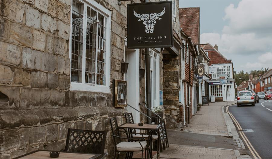 The Bull Inn sign above outdoor seating on a historic village street in Battle, lined with stone buildings and shops.