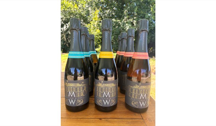 Bottles of Mountfield Winery sparkling wines with coloured labels displayed on a wooden table outdoors.
