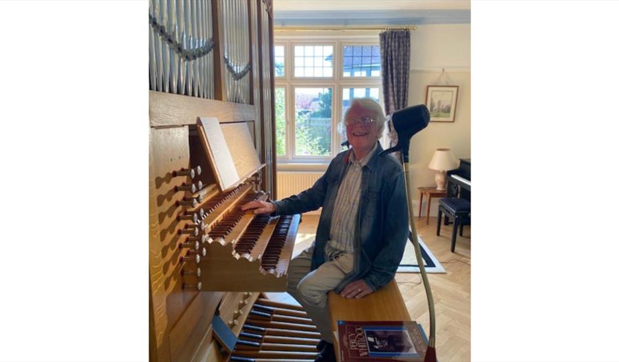 Person seated at a pipe organ at St Nicolas, Pevensey, with keys, pedals and stops visible in a bright room.
