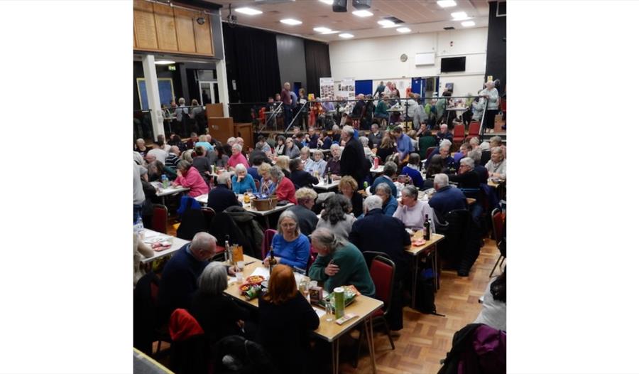 Large community gathering in a hall, with groups seated at tables enjoying food and conversation.