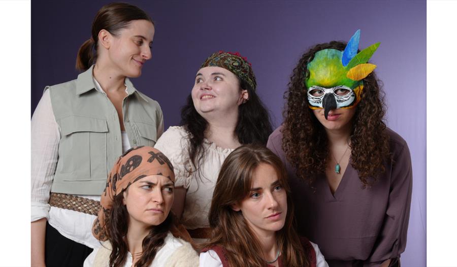 Studio photo of five costumed performers. The rear row features a smiling person in a light utility vest, a person with a patterned headscarf smiling