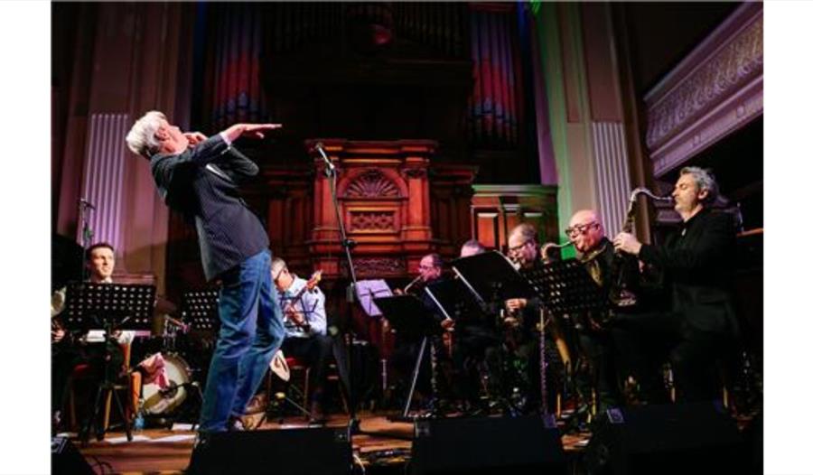Musician performing energetically on stage with a live band in a grand hall featuring ornate wooden panels and organ pipes.