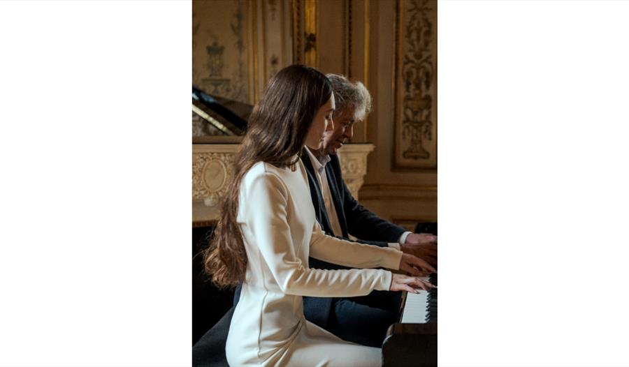 Two musicians seated at a grand piano, performing a duet in an ornate, gilded room with classical décor.