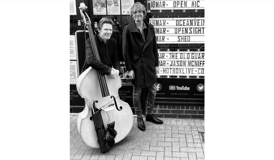 Two people standing outside a venue with a large double bass, next to a sign listing upcoming music events.