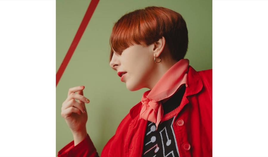 Image of Laura Fell with short auburn hair wearing a red jacket, pink scarf and patterned top against a green wall.