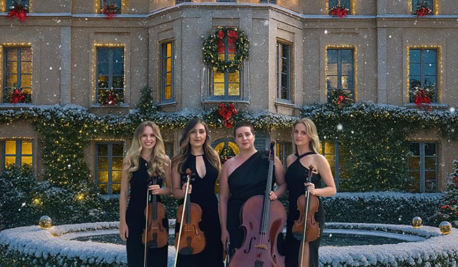 Image shows four women musicians standing outdoors in front of a grand mansion decorated for Christmas. They each hold string instruments — two violin