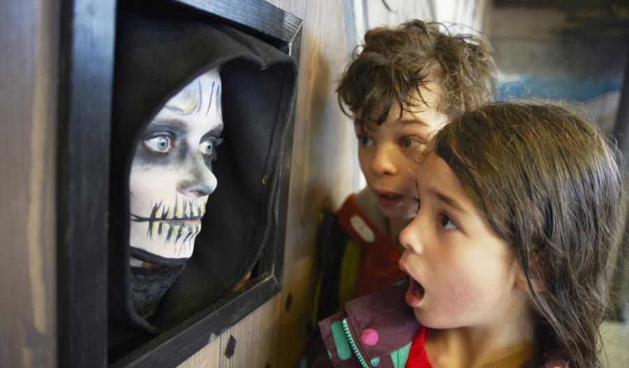 Two shocked looking children to the right look at wall on the left. Wall has a black frame with a human face painted like a skeleton.