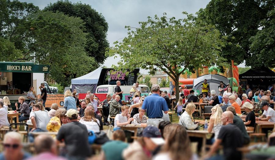 a landscape view of a country festivals. Crowds of people on picnic benches with food bars and a tent in the background.