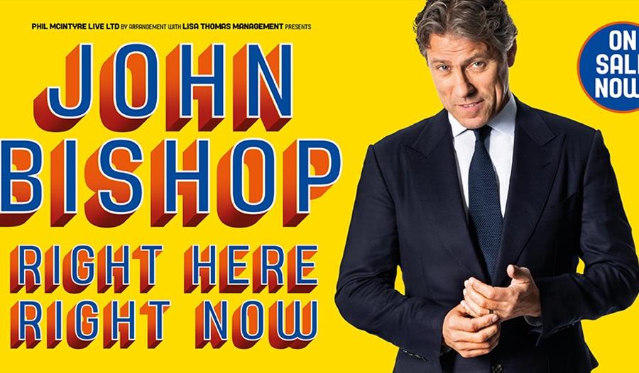 A poster with a yellow background. To the right a white man in a suite. Left says John Bishop. Right here right now.