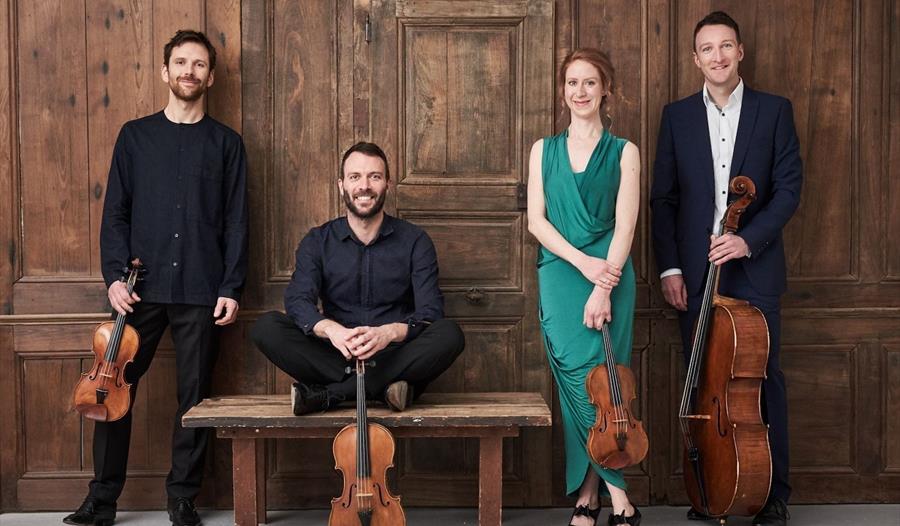 a photograph of four musicians standing in a row against wooden panelled wall. three men in dark suits, one sitting crossed legged on bench, others st