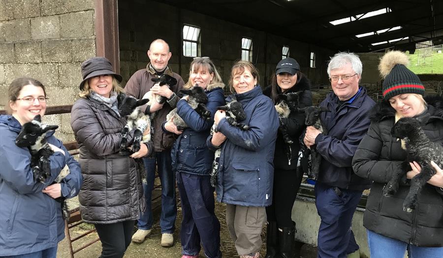 A group of eight adults in coats outside an open barn door, each holding a lamb in their arms.