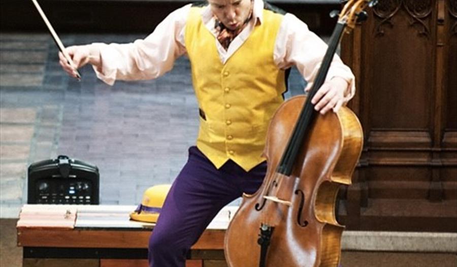 A man in a yellow waistcoat and purple trousers plays a cello on stage. For a Rye Creative Centre event, Rother.