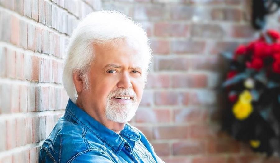 a photograph of Merrill Osmond against a brick wall, flowers in background. A man with white hair and beard.