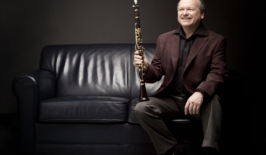 A photograph of a man sat on a black sofa with a clarinet resting on knee. Black background, man wears black suit.