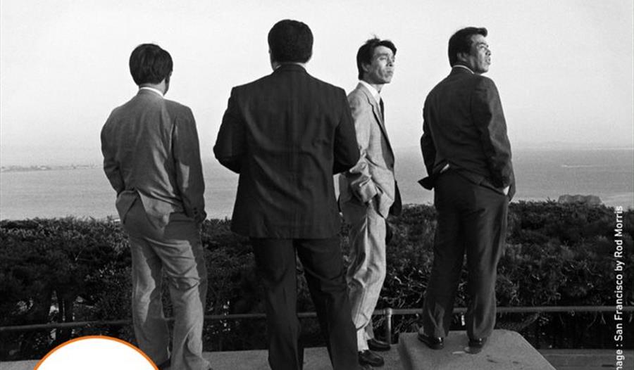 A poster for the Photohastings Summer exhibition. Image features four men in suits looking out to sea.