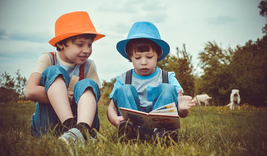 a photograph of two children sat on grass, one wearing an orange trilby hat, the other blue. Boy on right reads a book