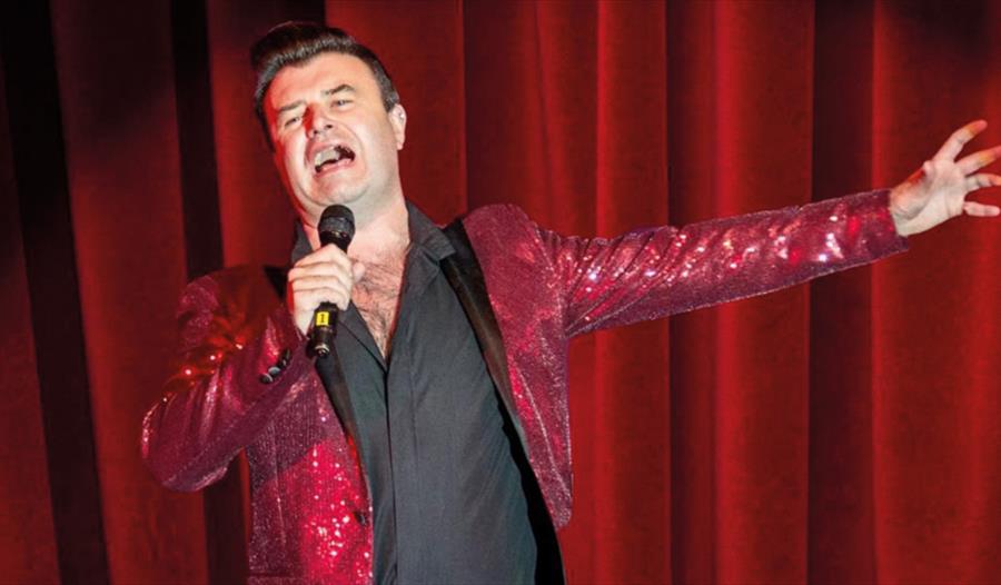 a man singing wearing a shiny red jacket against a red curtain.