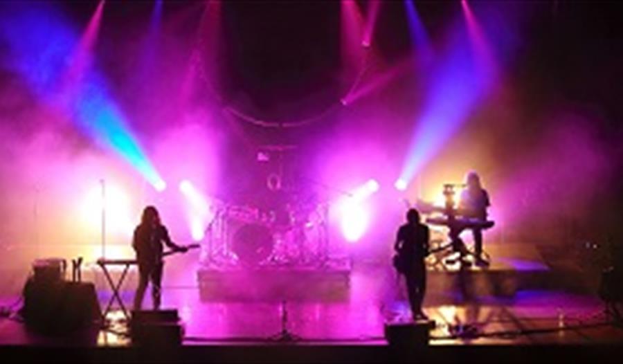 a photograph of think floyd on stage. Silhouette of band against bright pink, yellow and blue stage lights.