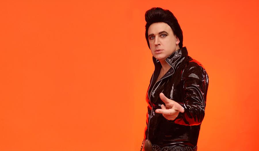 man standing before plain bright orange background. white man in leather jacket and high brown hair, like Elvis Presley.
